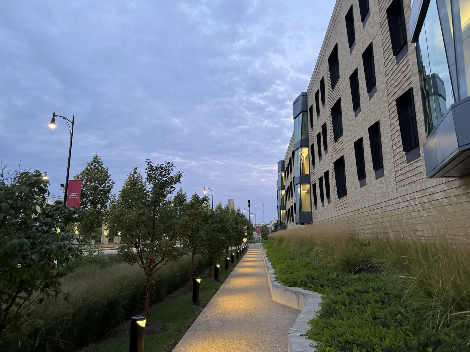 A lit campus path at dawn beside a brick building, under a pale cloudy sky